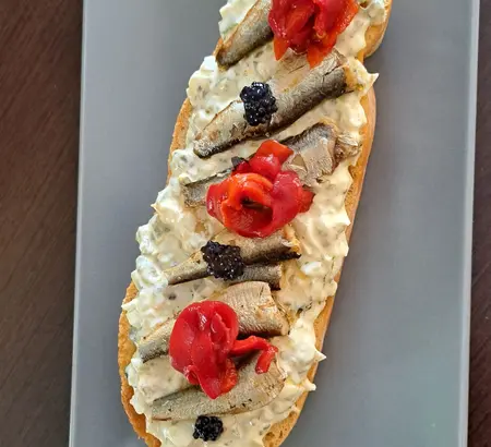 Tosta alargada con sardinas, crema, pimiento rojo y perlas negras